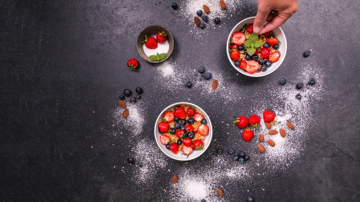 Fresh strawberries, blueberries, almonds in bowls on black table with hand arranging.